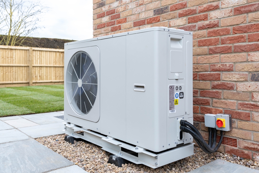 A white heat pump unit is installed outside a brick house in Indiana on a gravel bed, with cables connected to a nearby power switch.