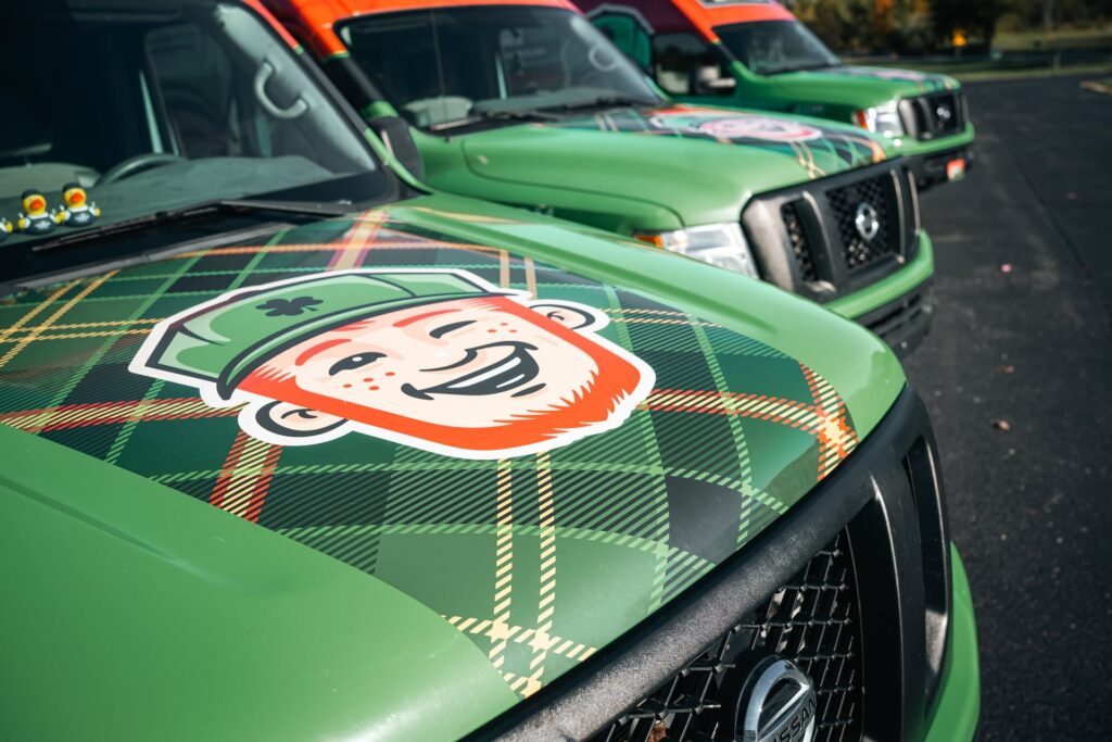 Green trucks parked in a row in Franklin IN, each with a plaid pattern and the cartoon face of a smiling red-headed character in a green cap on the hood—perfectly representing local ductwork services.